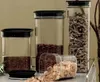 Several clear plastic food storage containers with black lids are displayed on a shelf. One container is filled with pasta, another with red beans, and a third with small seeds. A fourth, smaller container is tipped over, spilling brown sugar crystals. Anteprima dell’immagine prodotto