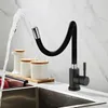 A black kitchen faucet with a flexible spout is shown in use, with water spraying from it into a sink. Several white ceramic containers with wooden lids are placed on the counter behind the faucet. Náhľad obrázku produktu