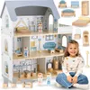 A young girl is sitting in front of a large wooden dollhouse, smiling at the camera. The dollhouse is fully furnished with miniature furniture and accessories. Produktbild-Vorschau
