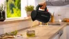 A person is pouring hot water from a black electric kettle into a glass teapot on a kitchen counter. Anteprima dell’immagine prodotto