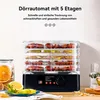 A food dehydrator with multiple trays filled with dried fruits and vegetables, placed on a wooden surface with some fresh fruit and a jar of dried oranges nearby. Produktbild-Vorschau