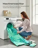 A woman is placing a large, green blanket with a tropical leaf pattern into a washing machine. Produktbild-Vorschau