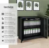 A black metal filing cabinet filled with binders is shown in a room with green walls. To the left of the cabinet is a panel with text and icons describing its features. Three framed botanical prints hang on the wall above the cabinet. Produktbild-Vorschau