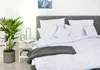 A neatly made bed with white bedding and a gray throw blanket, next to a nightstand with a plant and lamp. Podgląd zdjęcia produktu