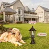 A golden retriever dog lies on a green lawn in front of a large house. A garden lamp is placed in the foreground with dimensions indicated. Produktbild-Vorschau