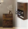 A shoe cabinet with two flip-out drawers is shown in a room setting, with a vase and plant on top of a similar cabinet to the left. The shoe cabinet is open, displaying shoes inside. Náhled obrázku produktu