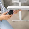 A person's hands are shown attaching a black caster wheel to the white leg of a height-adjustable desk. Produktbild-Vorschau