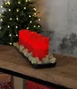 Four red LED candles on a black tray with moss, placed on a wooden shelf in front of a decorated Christmas tree. Produktbild-Vorschau
