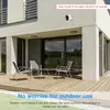 A security camera is mounted on the exterior wall of a modern house, with a patio and outdoor furniture visible below. The image highlights the camera's suitability for outdoor use. Podgląd zdjęcia produktu