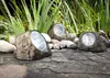 Three solar-powered rock lights are placed on a stone surface in front of lush green plants. Produktbild-Vorschau