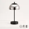 A modern black and silver table lamp with a dome-shaped shade, placed on a white surface against a white background. Produktbild-Vorschau
