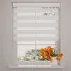 A white roller blind with horizontal stripes of opaque and sheer fabric is shown installed on a window. Orange tulips in a white vase sit on the windowsill. Produktbild-Vorschau