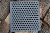 A square metal grate with many holes rests on a pile of chopped wood. Produktbild-Vorschau
