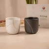 Two kintsugi cups, one white with gold repair lines and one dark gray with silver repair lines, are placed on a light beige surface. A box with the brand name "CHIYU" and "Kintsugi" is visible in the background. Podgląd zdjęcia produktu