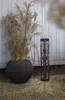A black planter with dried ornamental grass and a black lantern stand on a gravel path in front of a white wooden wall. Produktbild-Vorschau