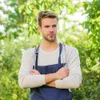 A man wearing an apron and holding barbecue tools, standing outdoors with a blurred green background. Anteprima dell’immagine prodotto