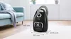 A black Bosch vacuum cleaner is shown in a living room setting next to a blue couch. Produktbild-Vorschau