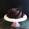 A dark chocolate bundt cake on a white cake stand with a rose gold base, set against a dark background. Podgląd zdjęcia produktu