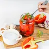A hand is using a fork to take a piece of pickled red pepper from a glass jar. Other ingredients like garlic and salt are also present on a wooden board. Produktbild-Vorschau