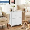 A white two-drawer nightstand is placed next to a beige armchair and a sofa, with a golden retriever lying on the floor in front of it. A vase of white flowers and a mug are on top of the nightstand. Produktbild-Vorschau