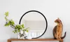A round mirror with a black frame is displayed on a white wall above a wooden shelf. A vase with flowers and a small white vase are on the shelf, along with a cat sitting to the right of the mirror. Produktbild-Vorschau
