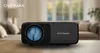 A black projector is placed on a wooden table in a blurred living room setting. Voir les images du produit