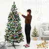A woman is decorating a Christmas tree with lights and ornaments in a room with another smaller Christmas tree and a sofa. Náhled obrázku produktu