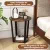 A round side table with two shelves, placed next to a bed. The table has a rustic brown wood finish and black metal legs. It is decorated with a lamp, books, and a small clock. Náhled obrázku produktu