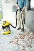 A person is using a yellow and black wet/dry vacuum cleaner to clean up fallen leaves on a gravel path next to a building. Produktbild-Vorschau