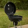 A black charcoal grill with the lid open, placed on a paved surface with grass in the background. Produktbild-Vorschau