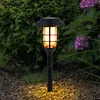 A solar-powered garden light with a flickering flame effect is placed in a garden bed with gravel and plants. Produktbild-Vorschau