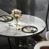 A round marble-topped table set for two, with a vase of pink roses, a drink, and place settings. It is raining, with water droplets on the table and surrounding area. Anteprima dell’immagine prodotto