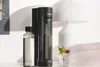 A black SODAPOP water carbonator sits next to a glass bottle of water and a glass of sparkling water with fruit and mint, with a white lamp in the background. Anteprima dell’immagine prodotto