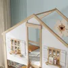 Close-up view of a children's house-shaped bunk bed with wooden accents and white walls. Features include windows with shutters and a small shelf with decorative items. Produktbild-Vorschau