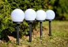 Four white globe-shaped solar garden lights are lined up in the grass in front of a green bush. Produktbild-Vorschau