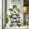 A black metal plant stand with multiple tiers and heart-shaped shelves, filled with various potted plants, is placed near a window with vertical blinds. The background shows a wall with framed artwork and a glimpse of a shelf with decorative items. Náhled obrázku produktu