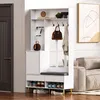 A white entryway organizer with a bench, shoe storage, and hooks, set against a neutral background with a door and a sofa visible.