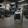 A heater is placed on the floor of a car repair shop, with a car lifted on a hydraulic lift in the background. Podgląd zdjęcia produktu