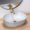 A close-up shot of a beige oval countertop sink with a gold faucet, set against a wooden surface and a circular mirror. Náhled obrázku produktu