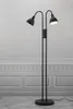 A black floor lamp with two adjustable heads stands against a white and gray paneled wall. Náhled obrázku produktu