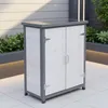 A white and gray wooden garden cabinet with two doors is shown outdoors on a tiled patio. Green plants are visible in the background. Produktbild-Vorschau