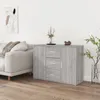 A light gray wooden sideboard with three drawers and two doors is shown in a living room setting next to a sofa. There is a vase of flowers and a decorative object on top of the sideboard. A framed abstract picture hangs on the wall. Produktbild-Vorschau