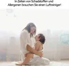 A mother and child are embracing in a bright, airy room, with abstract representations of airborne particles or viruses in the background. Produktbild-Vorschau