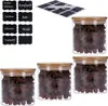 Four glass jars filled with raisins, each with a bamboo lid, are arranged on a white background. A set of black chalkboard-style labels is also visible in the upper left corner. Produktbild-Vorschau