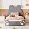 A child's bed with a bear-shaped headboard and paw prints on the footboard, set in a bedroom with bedside tables and a lamp. Produktbild-Vorschau