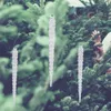Three clear acrylic icicle decorations hang in front of a blurred background of green Christmas trees, with a person's hands visible in the upper right corner, adjusting a tree branch. Podgląd zdjęcia produktu