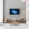 A modern living room setup featuring a Thomson TV displaying a galaxy image, mounted above a wooden shelf with electronic devices and decorative items. A coffee table and rug are in the foreground. Anteprima dell’immagine prodotto