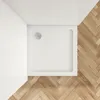 A top-down view of a white square shower tray with a drain in the corner, placed on a wooden floor with a herringbone pattern. Produktbild-Vorschau