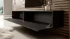 A modern, wall-mounted black TV stand with its two front doors open, revealing the interior. The stand is mounted on a slatted wooden wall. Decorative vases are visible on top of the stand. Voir les images du produit
