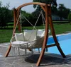A wooden swing chair with white cushions sits on a stone patio next to a swimming pool. Produktbild-Vorschau
